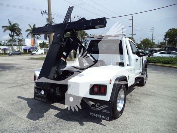 White Ford F-450 tow truck with a large black lifting apparatus mounted at the rear. Visible text includes a phone number on the rear fender and a large hitch mechanism.