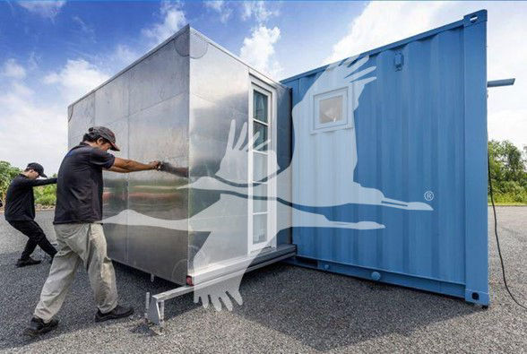 Two people move a modular trailer into a blue shipping container. The silver module has a door and small window. Clear sky with scattered clouds.