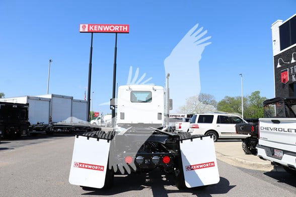 Kenworth T880 cab and chassis truck from rear view with large white mud flaps showing Kenworth logos; dealership lot in background.