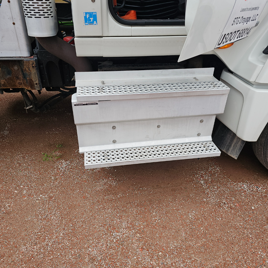 White perforated metal steps/footboards attached to a truck cab with gravel ground in background.