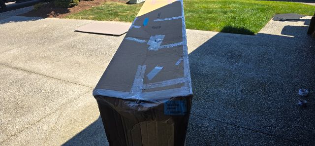 Boxed item on a pallet or stand with protective packaging and blue tape showing a long rectangular package.