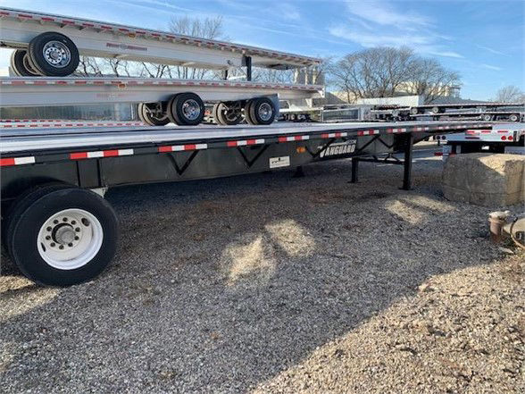 New Vanguard 48ft steel flatbed trailer showing long steel deck, triple-axle height, side rails, black undercarriage, white wheel with black tire, Vanguard logo on side.