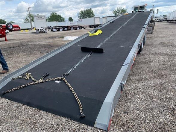 Container delivery trailer with a lowered drop deck, tan tandem axle, and heavy-duty chain and strap on the ramp.