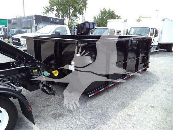 Isuzu NRR refuse truck body with large black dumpster-style container attached to hitch. No legible labels visible on container. Background shows other trucks and trailers.