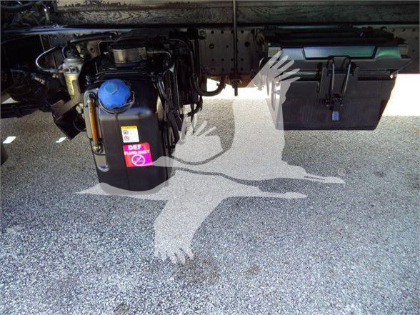 Underneath a truck showing a DEF fluid reservoir with blue cap and a pink label, plus a black battery box and other components.