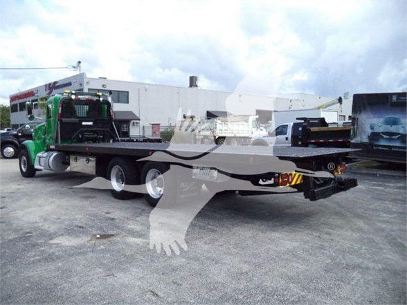 Tow truck with a long flatbed trailer, Peterbilt cab in dark green, and a black trailer. Visible text on the trailer rear includes hazard stripes and tail equipment. No legible model numbers or fluid labels readable.