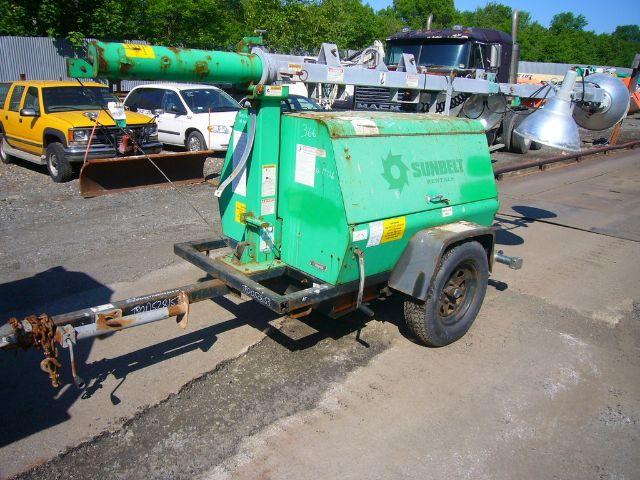 Green portable air compressor on a single-axle trailer with two wheels, attached hitch, and 'Sullair' branding visible on the side; other equipment and vehicles in the background.