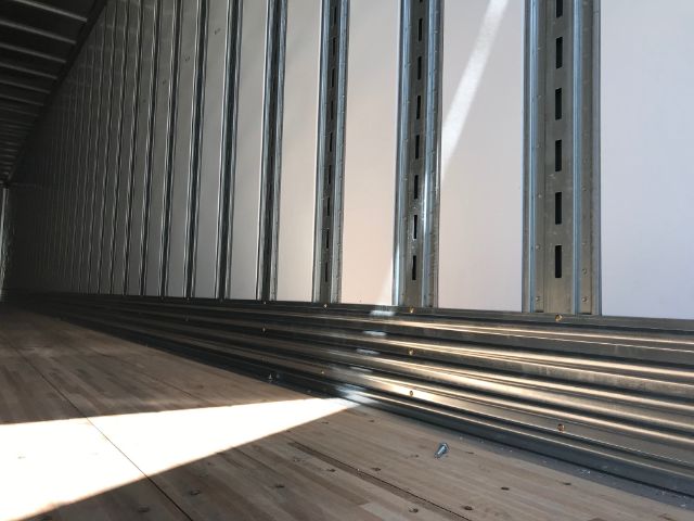 Interior of a cargo trailer with wooden floor and vertical steel wall studs, angled sunlight at the bottom.