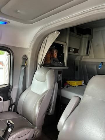Truck cab interior of a 2015 Peterbilt conventional sleeper showing driver's seat and passenger seat with a small bunk area and overhead storage.