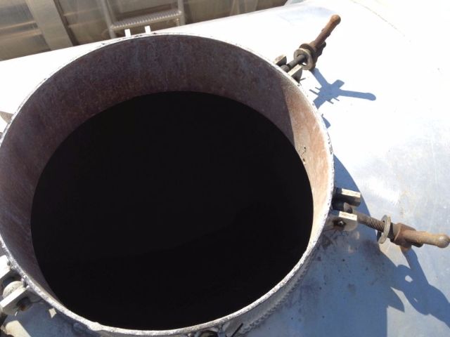 Open circular open hatch of a vacuum tank with rusted rim and flanges, showing interior darkness; exterior light gray metal surface with clamps and bolts.