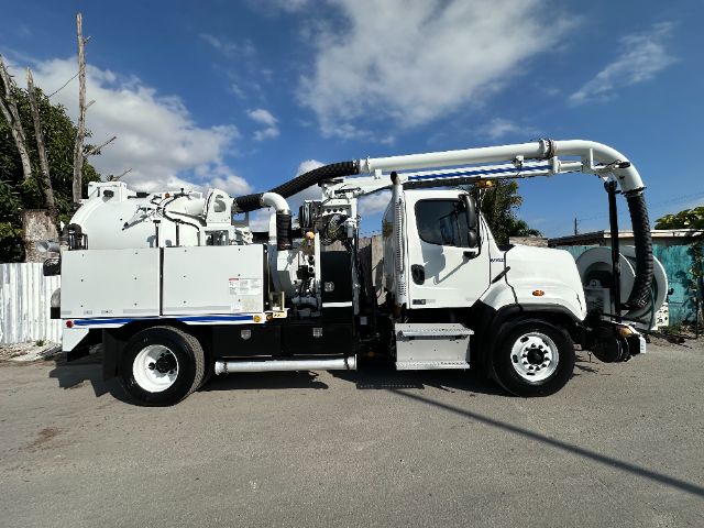 White Freightliner 108SD vacuum truck with long vacuum hose, side compartments, and mounted equipment; truck cab visible; blue sky.