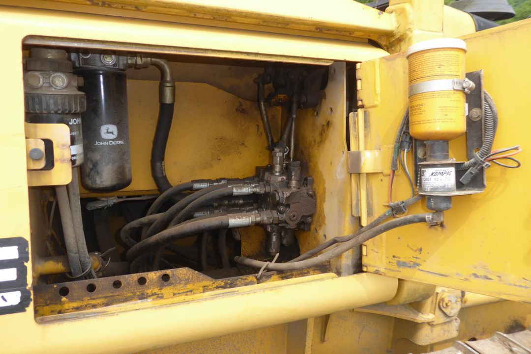 John Deere hydraulic reservoir and hoses inside a yellow Dozer cab area, with sheet metal firewall and black hydraulic lines, filter canister, and orange fluid reservoir on the right.