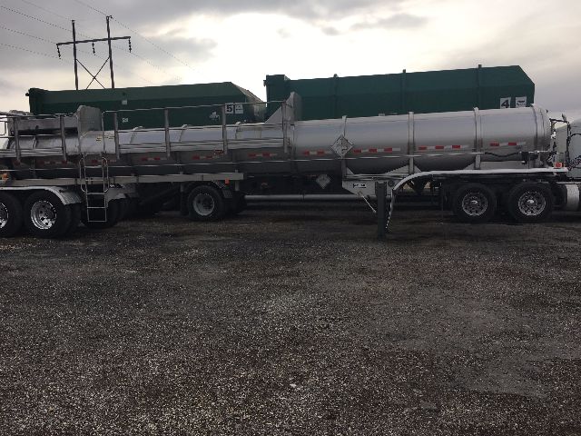 Trailer with large cylindrical tanker and green storage tanks in background. No legible text visible on the tanks or trailer.