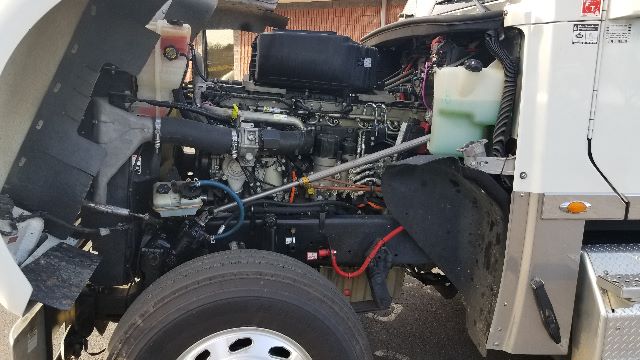 Truck engine bay of a 2013 Freightliner Coronado 132 showing exposed diesel engine, hoses, wiring, and front wheel with surrounding components.