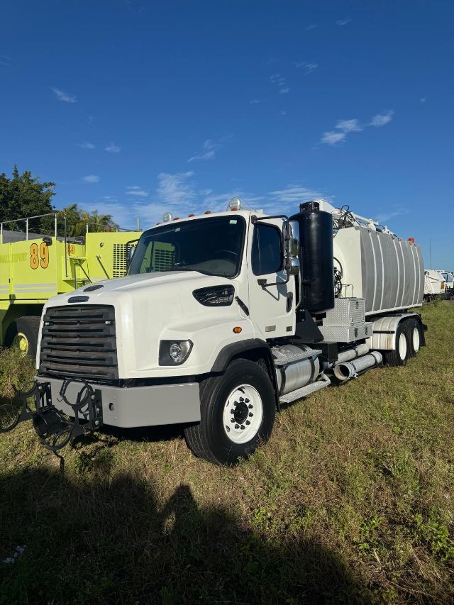 White Freightliner 114SD vacuum truck with large cylindrical tank on dual rear axles, black exhaust stack, front grille, and steps; clear blue sky.