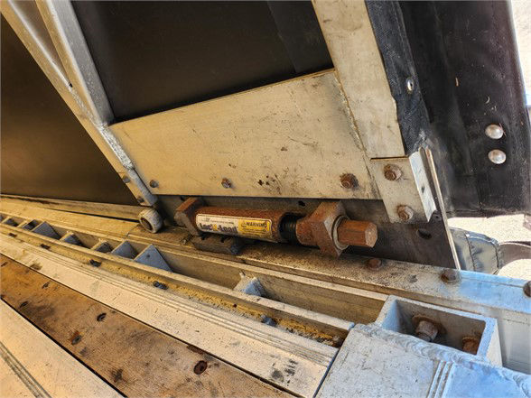 Trailer component with hydraulic latch/hinge mechanism and rusted hardware on a Manac curtainside trailer impact area.