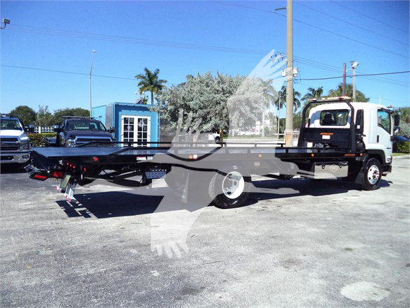 Isuzu FTR tow truck chassis with long flatbed seen in lot; white cab, black flatbed, rear equipment, blue building and palm trees in background.