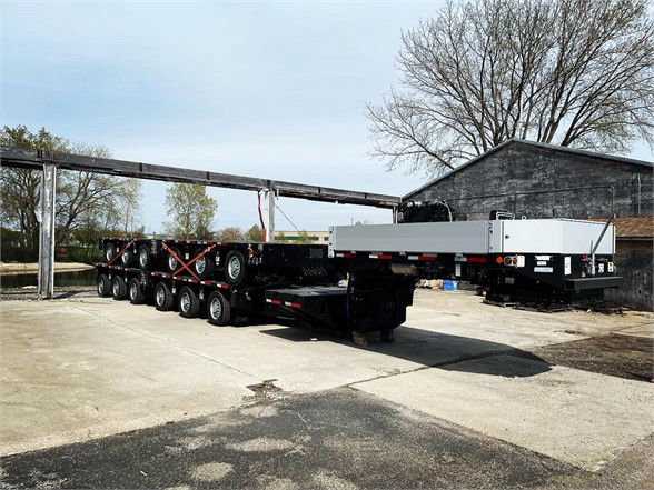 A large multi-axle trailer, Double Drop Deck, with a long flatbed and multiple wheels, parked on pavement near a building.