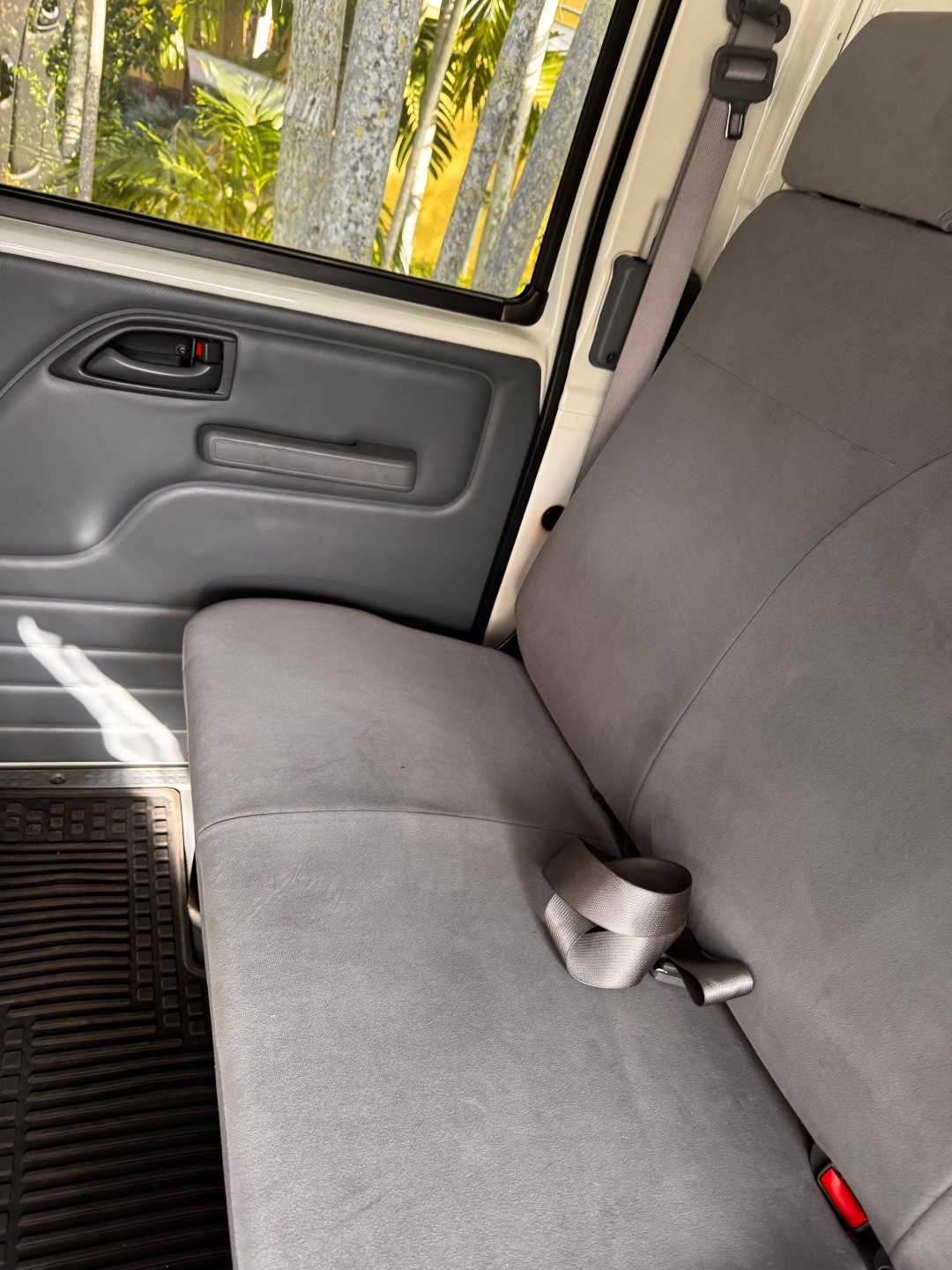 Isuzu NQR interior: front passenger-side door panel with manual window switch, gray upholstery, seat belt buckles, and gray bench seat.