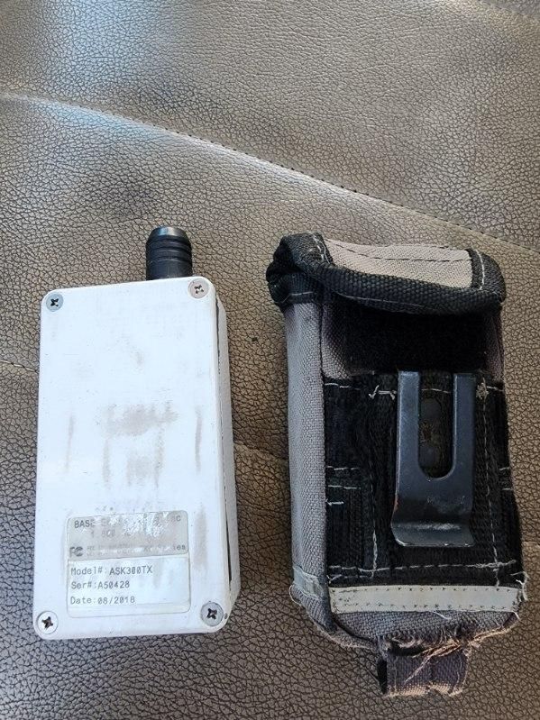 Trucking equipment image showing a small white electronic device with a black antenna and a black-wrapped pouch, both resting on a brown textured surface.