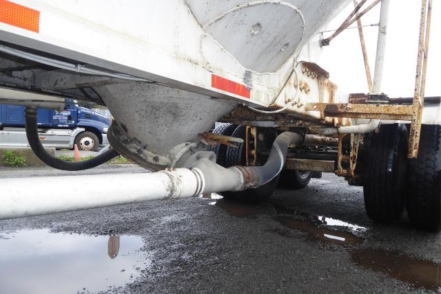 Bottom view of a Fruehauf dry bulk trailer with a large curved discharge pipe, rusted frame, and parked wheel in background. Puddles on wet pavement reflect the overhead. 
