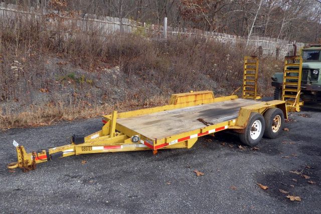 Trailer with flatbed and dual-axle on a gravel lot, yellow paint, ramps at rear, kingpin and hitch at front, weathered surface.
