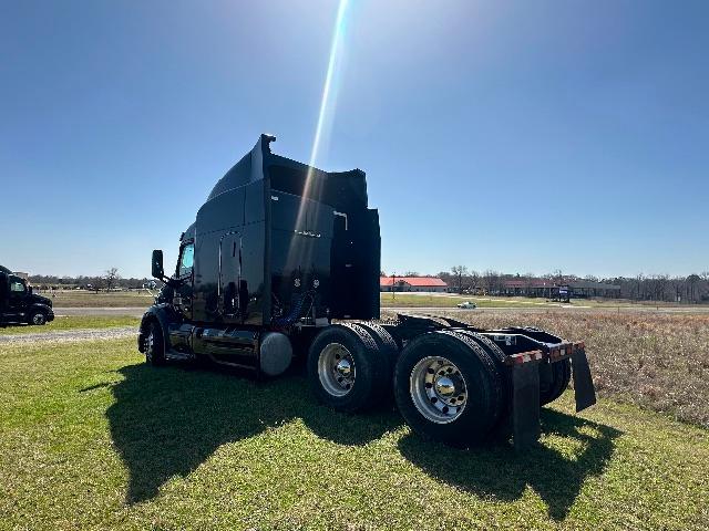 Truck tractor in a field, black Peterbilt 579 cab with sleeper, triple axles at rear, visible fuel tanks and wheels, clear sky.
