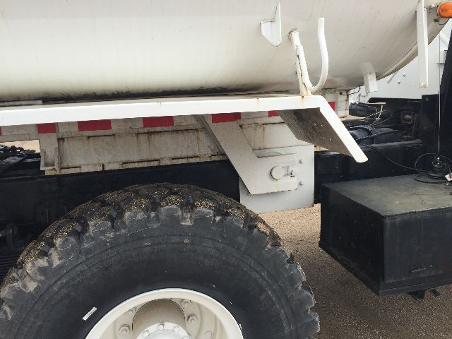 Stock wheel and undercarriage of a 1986 American General M923A1 military truck. Visible components include a large off-road tire with white wheel, chassis, and a boxed black tool/storage box. The tanker body sits above with a white support 