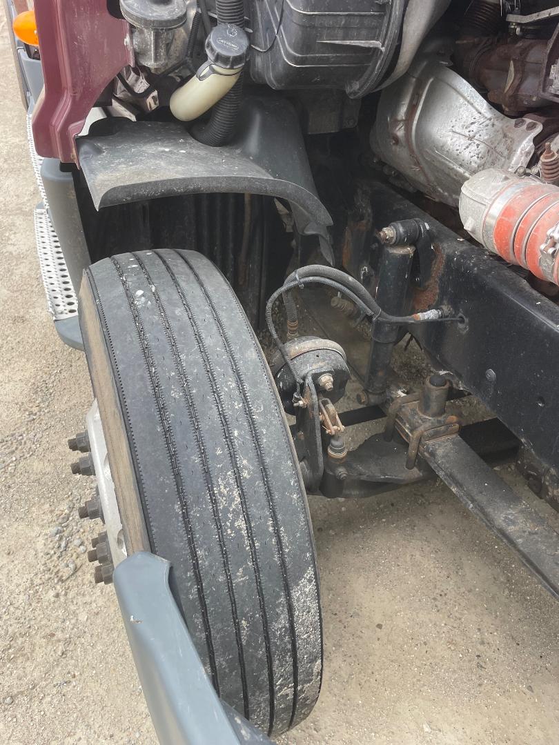 Front left wheel and suspension of a Freightliner Cascadia truck showing air brake line, dual tires, and engine bay components with a large black splash shield and exposed frame.