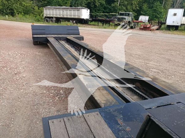 Lowboy trailer chassis showing long steel beams and deck cutouts, with gravel ground and other trailers in background.