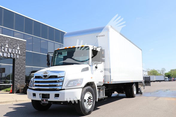 A white 2025 Hino L6 cab and chassis box truck parked outside a dealership. The truck has a large white box body and chrome grille; clear blue sky.