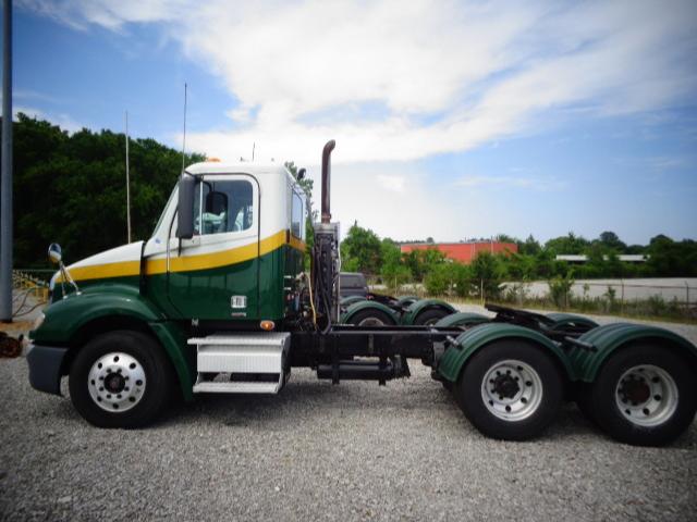 Freightliner Columbia daycab tractor showing green and yellow cab with side exhaust and dual rear axles on gravel lot.