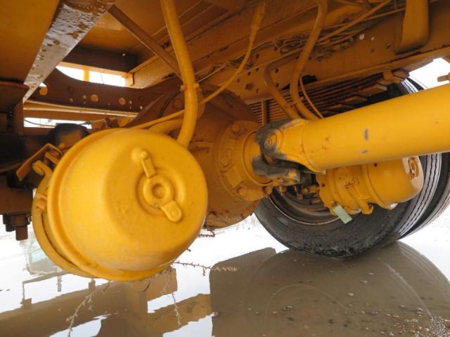 View of a yellow truck undercarriage showing a round reservoir and hydraulic components near the axle, with a large yellow driveshaft and wheel in the background.