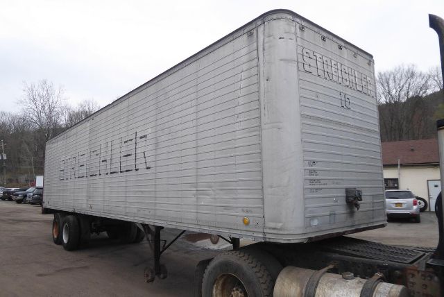 Trailer side and rear showing a long aluminum cargo trailer with ribbed siding, a raised front corner, and tires. Faded lettering on side and back, with wheels and hitch visible.