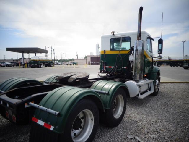Truck tractor in a yard, side and rear view showing fifth wheel, dual rear axles, green and white cab, exhaust stack, and fuel tanks.
