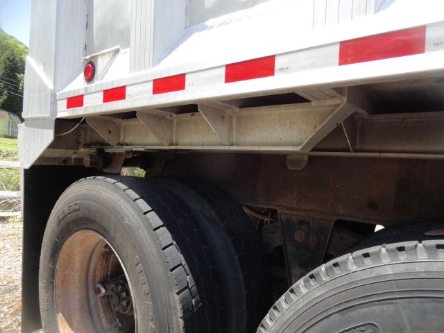 Trailer undercarriage showing dual rear wheels with worn tires and rust on suspension components; side of dump trailer with white and red reflective tape.