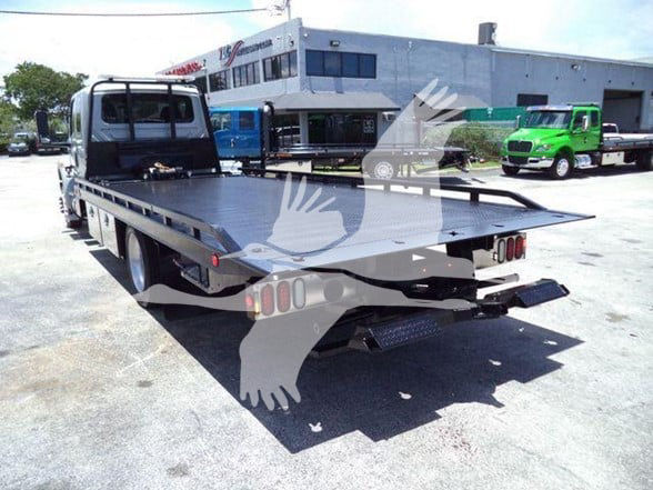 Flatbed tow truck with long metal bed and rear bumper lights, viewed from rear-left angle in a dealership lot.