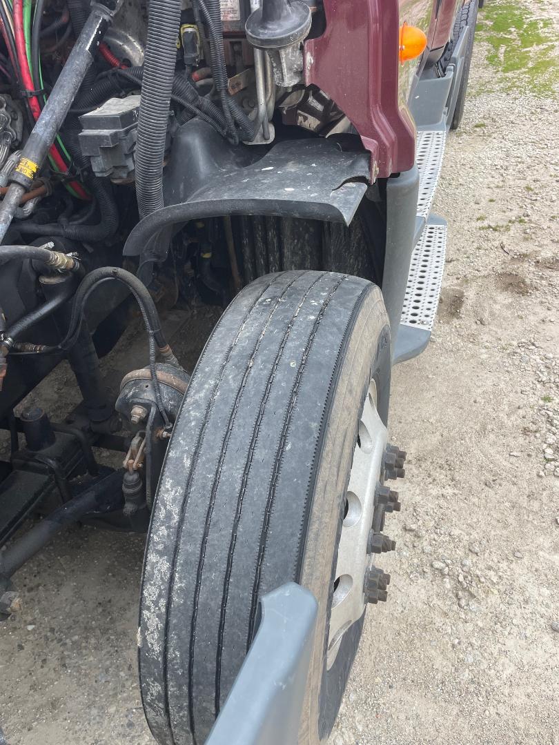 Front wheel of a Freightliner Cascadia 125 showing tire tread, wheel, and part of the fender and engine bay; visible hoses, wiring, and components.