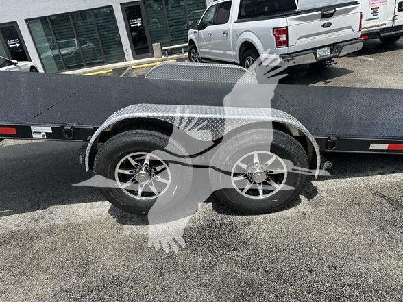 Flatbed trailer with dual axles and diamond plate fender showing two wheels; background includes a white pickup truck and a building with glass doors.