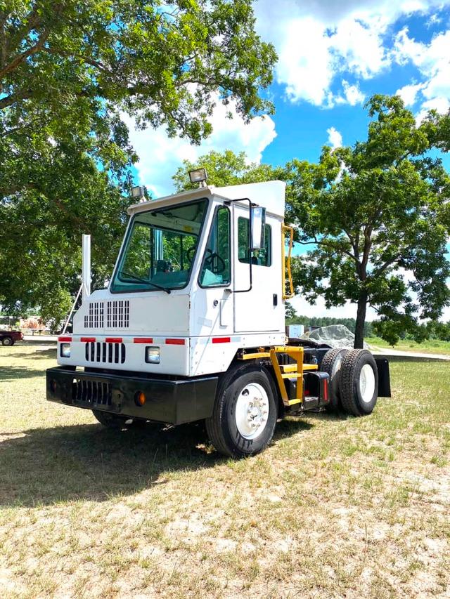 Truck yard-scale cab-over-engine style vehicle with white cab and yellow rear chassis. Front grille with two headlights, black bumper, and red reflective stripes. Large windows, flat open field and trees in background.