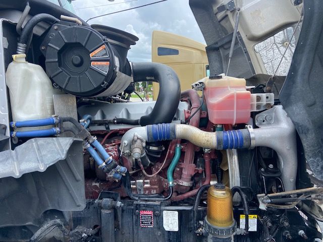 Engine bay of 2010 Mack CXU613 showing coolant reservoir, red coolant overflow bottle, yellow power steering/auxiliary reservoir, hoses, and engine components.