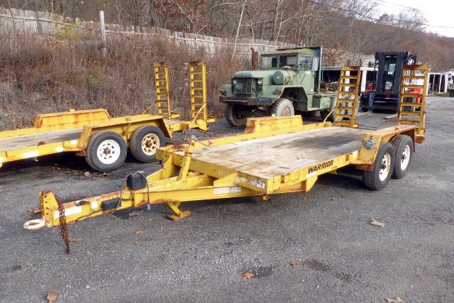 Trailer with yellow flatbed and ramps, labeled Warrior, in an outdoor lot with other trailers and a green military truck in background.