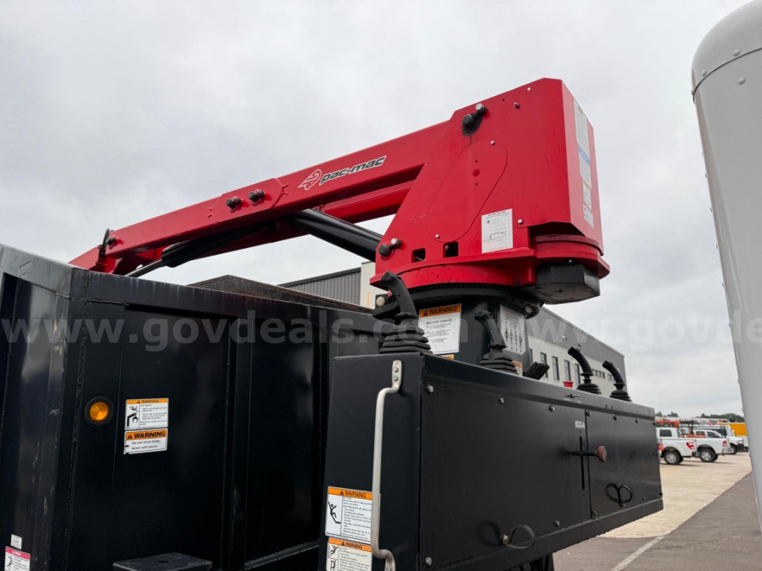 Peterbilt Pac-Mac KBF-20H grapple arm in red mounted on a black truck bed, with warning labels on the side and a grey overcast sky.