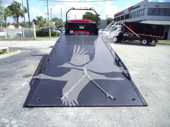 Tow truck flatbed with black diamond plate, two safety chains anchored on the deck, rear cab area, and a building in the background.