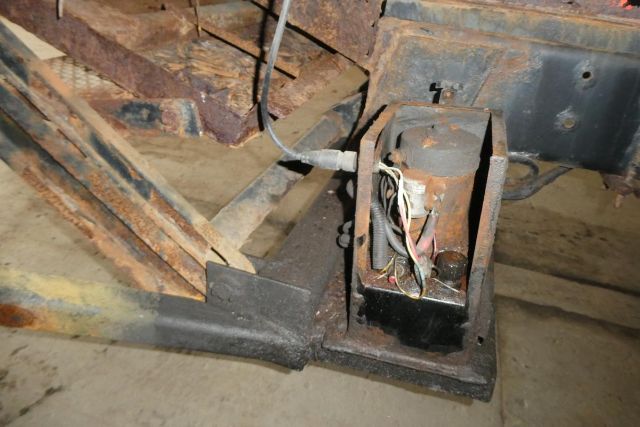 Truck chassis area with exposed electrical transformer-like component and wiring harness, rust and dirt on metal frame.
