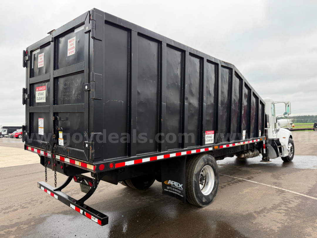 Truck side and rear of a black dump body grapple truck with safety decals, rear underride bar, and white cab visible on right.