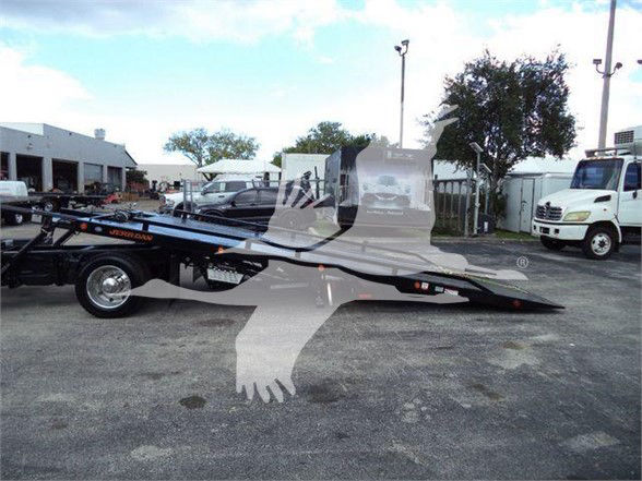 Tow truck trailer with a black hydraulic deck in a parking lot; faint watermark overlay. Text on the image includes small brand/logo on trailer and tires, but legible text is minimal.