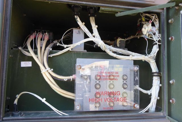 Truck electrical control panel with wiring harnesses and a high voltage warning label. Visible text: 'COVER MUST BE INSTALLED BEFORE OPERATING', 'WARNING HIGH VOLTAGE'. Various white cables enter a metal module with bolts.