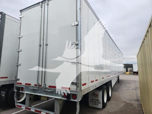 Trailer rear and side view of a long white van trailer with dual rear axles, reflective red/white tape, and doors; visible branding sticker on mud flap area.
