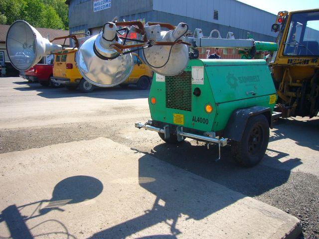 Green Terex AL4000 portable light tower/trailer with large floodlights and twin axle, mounted on green generator unit, in a yard with other equipment.